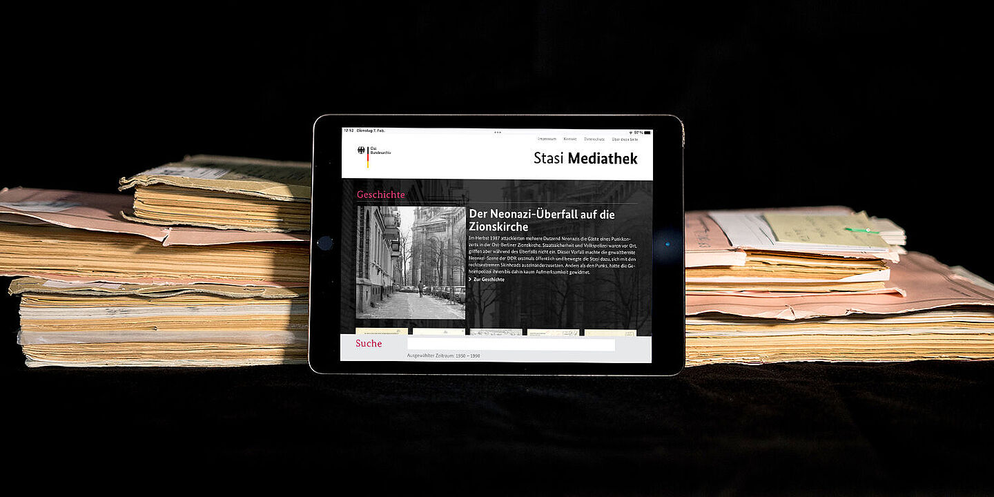 Das Bild zeigt die auf einem Tablet geöffnete Startseite der Stasi-Mediathek. Das Tablet lehnt an einigen Aken., Quelle:
            BStU / Mulders